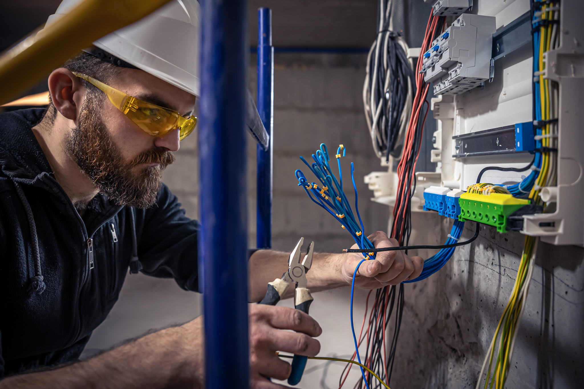 A bearded electrician, wearing a white hard hat and yellow safety glasses, is working with blue wiring using pliers. He is in front of an electrical panel with various colored cables and components inside a building under construction or renovation.