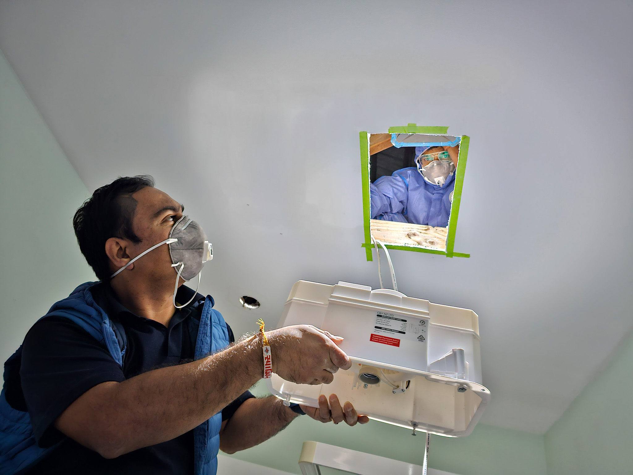 Two workers, both wearing face masks, collaborate on installing a ventilation system. One is inside a building holding the system up to the ceiling while the other looks down from an attic space through a small hole, guiding the installation.