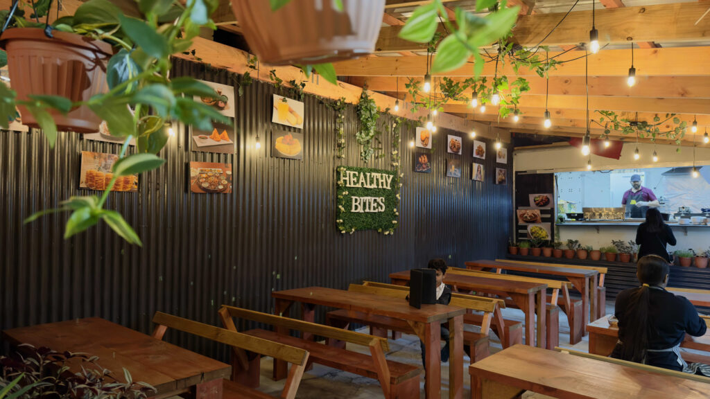 A cozy cafe with wooden tables and benches, decorated with hanging plants and string lights. A sign on the wall reads "HEALTHY BITES" surrounded by leafy vines. Customers are seated at tables, and a counter with food preparation is visible in the background.