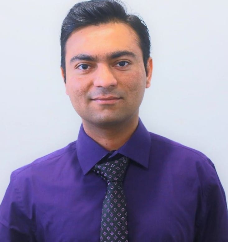 A man with short black hair is wearing a purple dress shirt and a black tie with a diamond pattern. He is facing forward against a plain light grey background.