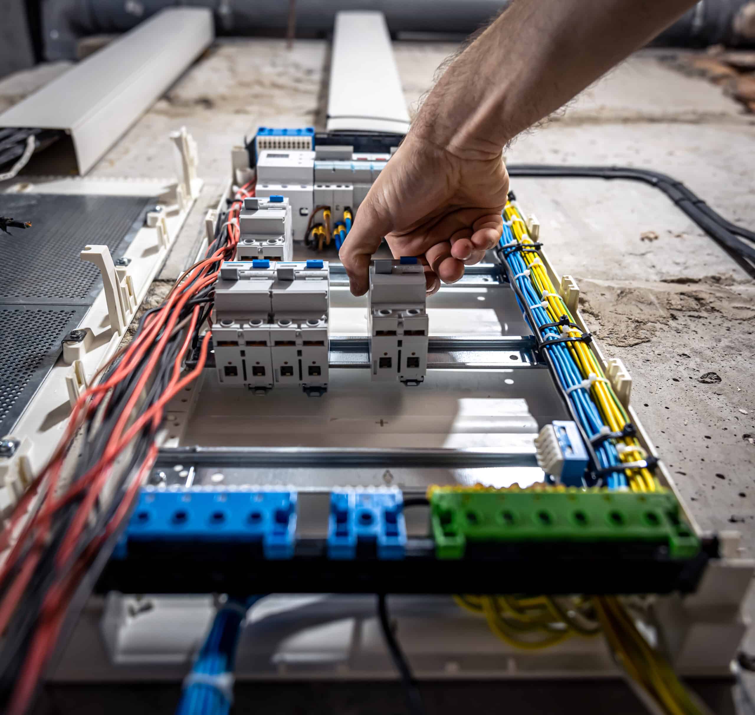 A person's hand adjusting circuit breakers inside an open electrical panel with various colorful wires organized around it. The scene appears to be part of an electrical installation or maintenance.