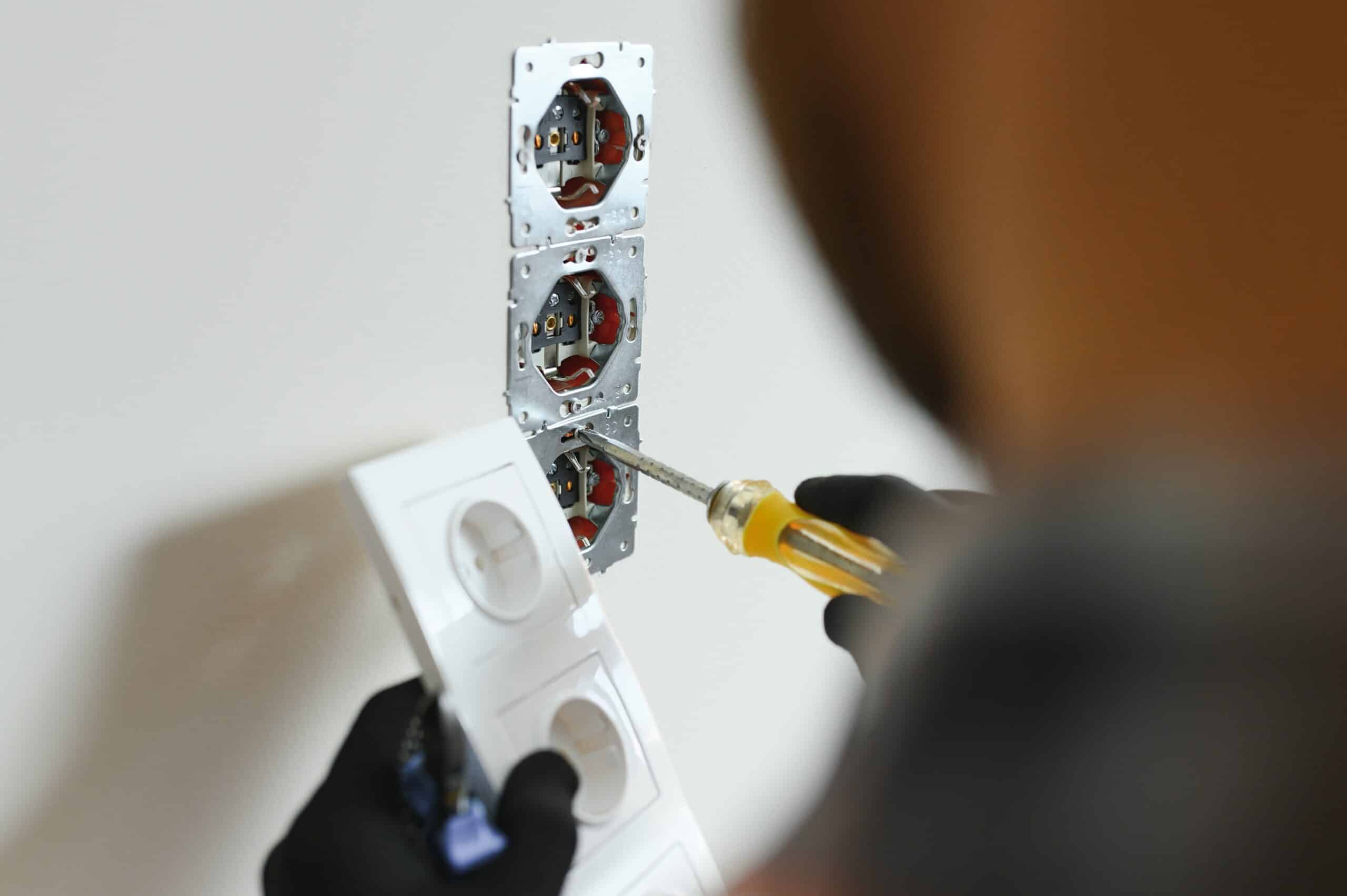 A person using a screwdriver to install or repair electrical outlets on a wall, with three outlet frames visible. The individual is wearing black gloves and appears to be following a set of instructions or guidelines.