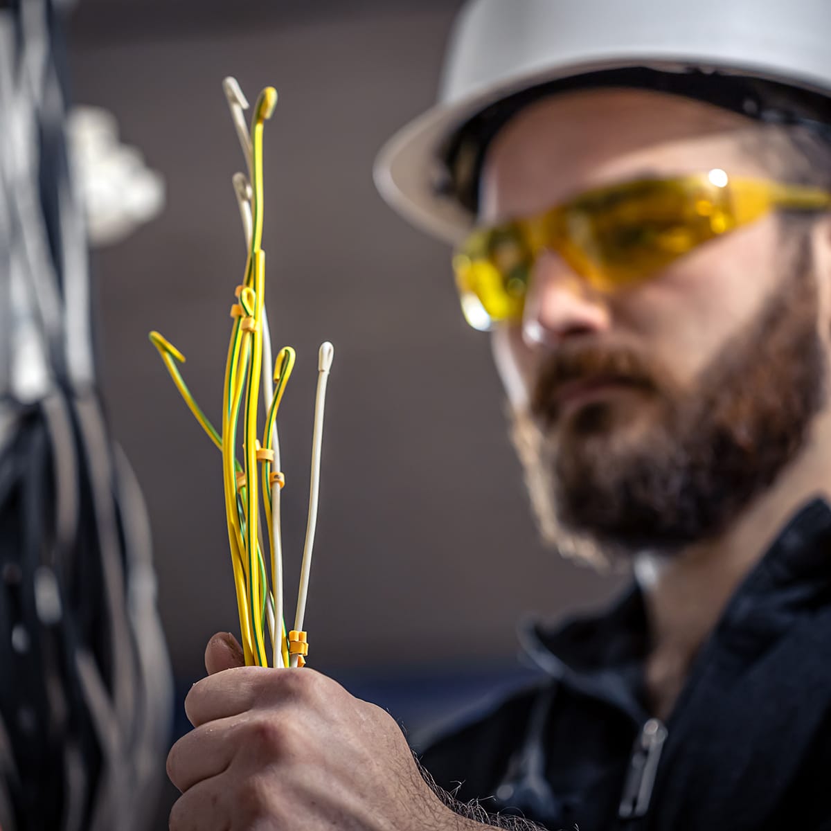 A person wearing a white hard hat and yellow safety glasses is holding several yellow and white wires. The background is slightly blurred, focusing on the wires in the person's hand.