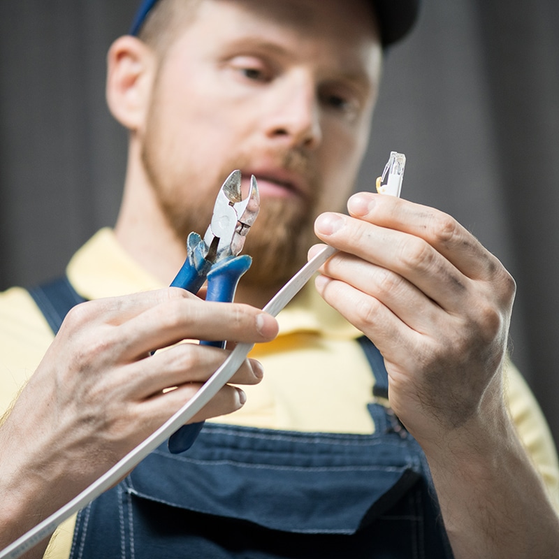 A person in overalls and a cap is holding a white cable in one hand and a pair of wire cutters in the other, appearing to prepare for cutting or stripping the cable. The background is blurred, focusing attention on the hands and tools.