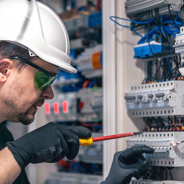 A person wearing a white hard hat, green safety glasses, and black gloves is using a red and yellow screwdriver to work on an electrical panel with numerous wires and components.
